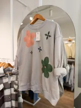 Buzo oversize color gris claro con cuello redondo, mangas largas y bordados de flores en tonos naranja, verde y azul. Confeccionado en tejido con interior de felpa.