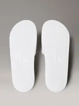 Ojotas tipo slide Calvin Klein Jeans, color blanco, con banda superior de lona de algodón que presenta el monograma de la marca en color negro. Posee suela de EVA con plantilla ergonómica moldeada.