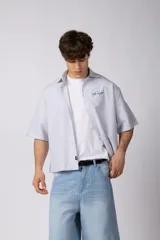 Camisa de manga corta de popelina rayada, con calce boxy y bordado sutil en el pecho. Presenta cierre frontal con botones y diseño abierto.