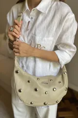 Cartera bandolera de cuero negro con diseño de media luna, decorada con tachas metálicas plateadas. Cuenta con correa regulable, cierre principal y bolsillo interior con forro.
