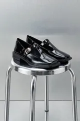 Mocasín estilo T-bar color negro en charol, con punta cuadrada, tira en el empeine con hebilla dorada y suela plana.