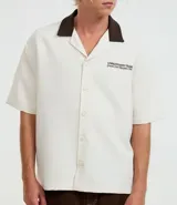 Camisa de manga corta, corte boxy, color crema, con cuello inglés y botones frontales. Presenta un bordado pequeño en el pecho izquierdo con texto en color marrón oscuro.