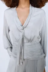 Camisa de escote en pico con lazada frontal en el mismo tejido, manga larga con puños y bajo drapeado. Presenta un acabado satinado y cierre lateral con cremallera oculta.