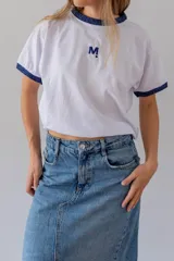 Remera blanca de algodón con cuello redondo y mangas cortas con puños de lurex azul. Tiene un bordado con la letra M en el pecho.