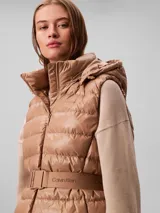 Chaleco puffer acolchado color tostado de la marca Calvin Klein, con diseño sin mangas, cuello alto y capucha desmontable. Cuenta con cierre frontal de cremallera y un cinturón elástico ajustable con hebilla de clip con el logo de la marca.