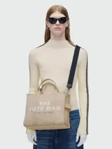 Bolso tote mediano de lona color celeste con estampado frontal del logo "The Tote Bag" en blanco y el nombre de la marca "Marc Jacobs" en la parte inferior. Tiene doble asa de mano y correa de hombro ajustable y extraíble.