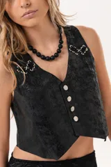 Chaleco de cuero negro con acabado efecto envejecido, escote en V y cierre frontal de botones metálicos. Presenta detalles de ojalillos decorativos en los hombros.