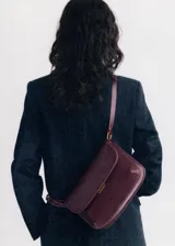 Bolso tipo bandolera de tamaño pequeño con acabado efecto piel en color bordó. Presenta una solapa frontal con cierre metálico a presión, costuras visibles en contraste y una correa larga ajustable con herrajes dorados.