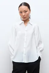 Camisa blanca de manga larga con puños abotonados y fruncidos, hombros fruncidos y cierre frontal con botones.