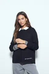 Remera de manga larga negra con cuello camisero blanco y puños a rayas blancas y negras. Presenta un diseño de corte holgado con un pequeño estampado de texto en la parte inferior frontal.