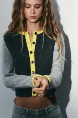 Chaqueta de punto con diseño color block, cuello redondo y manga larga. Presenta cuerpo en color gris oscuro, mangas en gris claro y detalles de ribetes en color amarillo vibrante. Cierre frontal con botones oscuros.