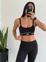Conjunto deportivo compuesto por un top de breteles finos con logo estampado y un pantalón largo de corte acampanado con aberturas laterales en el bajo. Ambas prendas son de color negro con detalles de costuras blancas a contraste.