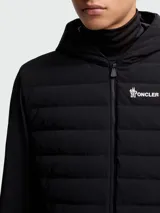 Chaqueta híbrida Moncler Grenoble negra con cuerpo acolchado y mangas de tejido liso. Presenta capucha fija, cierre frontal y logo de Moncler Grenoble en el pecho izquierdo.