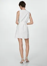 Vestido corto de color blanco con cuello en pico y diseño sin mangas. Presenta un corte recto, tirantes anchos y dos bolsillos de solapa en la parte delantera. Incluye un cinturón ajustable con hebilla metálica en la cintura.
