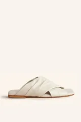 Sandalia tipo slide de cuero vacuno color blanco, con diseño de tiras cruzadas y fruncidas. Presenta una puntera cuadrada y una suela plana de cuero con un pequeño taco de 2 cm.