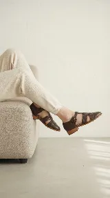 Zapato tipo cangrejera de cuero color marrón oscuro, con diseño de tiras entrelazadas, pulsera con hebilla ajustable al tobillo y taco bajo de madera.