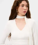 Blusa entallada de microfibra color blanco, con escote en V, mangas largas y detalle de frunces en los laterales. Presenta un lazo integrado en el cuello que permite anudarlo, aportando un toque sofisticado al diseño.