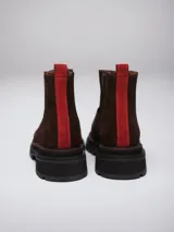 Botineta de gamuza marrón con detalles en cuero liso al tono. Presenta una suela track negra con costuras en contraste color rojo y una tira vertical roja en el talón.
