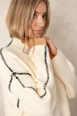 Buzo de punto acanalado color beige, con cuello alto, corte oversize y mangas largas. Presenta costuras decorativas en color negro que recorren los hombros y las mangas.