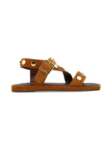 Sandalia plana de cuero gamuza en color camel, con tiras decoradas con tachas metálicas doradas y hebilla metálica de ajuste lateral.