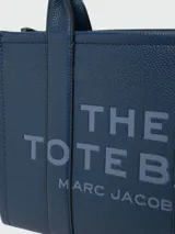 Cartera tipo tote de cuero granulado color tostado, con diseño estructurado y rectangular. Presenta doble asa superior fija y una correa larga ajustable y extraíble. En el frente, destaca el logo estampado con la inscripción 'THE TOTE BAG MARC JACOBS'.