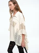 Camisa de corte oversize en color crudo, confeccionada con una mezcla de texturas que incluye paneles de encaje, detalles bordados y aplicaciones de tachas metálicas. Presenta mangas amplias tipo murciélago, cuello clásico y cierre frontal con botones.
