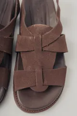 Sandalias planas de gamuza color marrón, con diseño de tiras cruzadas en el empeine y cierre con hebilla metálica dorada en el tobillo. Cuenta con plantilla de cuero y suela de goma con textura.