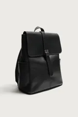 Mochila de diseño minimalista en color negro, confeccionada en material sintético con acabado liso. Presenta una solapa frontal con cierre de hebilla decorativa y sistema de imán, compartimento principal con cremallera, bolsillos laterales externos, asa superior de mano y correas de hombro ajustables y acolchadas.