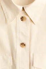 Camisa de estilo cargo color beige, confeccionada en mezcla de lino y algodón. Presenta cuello clásico, cierre frontal con botones, bolsillos superiores con solapa, charreteras en los hombros y mangas largas con puños abotonados.