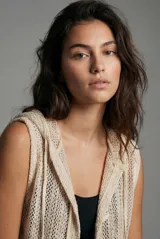 Chaleco de punto calado color beige, sin mangas, con capucha y bolsillos delanteros.