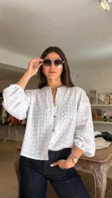 Camisa blanca de broderie con cuello redondo, cierre frontal con botones de madera y mangas largas abullonadas con puños elásticos.