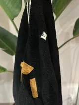 Buzo negro tipo corderito con cuello alto y cierre parcial con cremallera. Tiene un bolsillo tipo canguro con apliques de pana color mostaza y un parche con letras en el pecho. El ruedo y los puños tienen ribete de color.