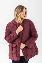 Chaqueta acolchada de corte oversize en color bordó, con diseño de costuras onduladas, cuello redondo y cierre frontal con botones a presión. Presenta bolsillos frontales tipo cargo con solapa y detalle de tiras.