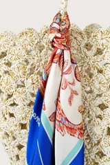 Pañuelo cuadrado de tejido ligero con estampado floral y botánico en tonos cálidos sobre fondo crema, enmarcado por un borde ancho en color azul vibrante con detalles de líneas blancas.