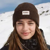 Gorro tipo beanie de tejido acanalado en color marrón, con parche rectangular frontal que incluye el logo de la marca y coordenadas geográficas.