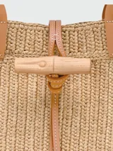 Bolso tipo shopper de gran tamaño, confeccionado en rafia natural con detalles de cuero en tono beige claro. Presenta asas superiores de cuero, cierre de broche tipo toggle con tira de cuero y estructura rígida.