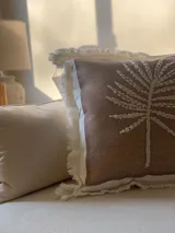 Almohadón cuadrado color marrón claro con bordado de hilo color crema con diseño de rayos de sol. Tiene borde blanco con flecos.