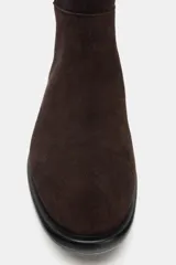 Bota de caña alta confeccionada en serraje, con diseño de estilo hípico. Presenta detalle de pespuntes verticales, tira decorativa con hebilla metálica en la parte superior de la caña y puntera redonda. Cuenta con suela plana de 2,5 cm y sistema de calce tipo pull-on.