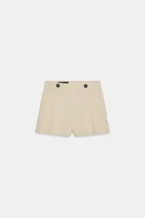 Short de tiro alto color beige, con detalle de pliegues frontales y aplicación de dos botones oscuros en la cintura. Cuenta con trabillas para cinto y bolsillos laterales.