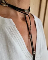 Collar tipo corbatín confeccionado en tiento de cuero negro, con detalles de cuentas metálicas plateadas distribuidas a lo largo de las tiras.