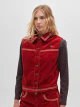 Chaleco de pana negro con cierre frontal de botones metálicos dorados. Presenta detalles de costuras en contraste en el cuello, cartera y canesú, e incluye un pequeño parche con forma de bandera en el pecho.