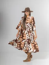 Vestido largo de manga tres cuartos, con cuello camisero y botones frontales. Presenta un estampado abstracto en tonos marrón oscuro, beige y blanco. El calce es amplio y tiene un ruedo con volado.