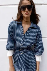 Vestido midi azul marino con rayas blancas, cuello solapa, escote en V y cierre frontal con botones. Cuenta con mangas abullonadas por debajo del codo con puños anchos a contraste, cinturón ajustable del mismo tejido con hebilla metálica y falda evasé.