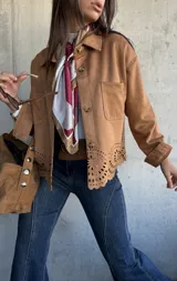 Chaqueta de gamuza color camel con cierre frontal de botones, cuello clásico y detalle de calados decorativos en el ruedo. Presenta bolsillos frontales y mangas largas con puños.