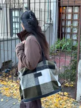 Bolso tipo tote de gran capacidad con diseño de cuadros en tonos verde oliva, blanco y negro. Presenta detalles en cuero negro en la base y en las correas de hombro, además de un parche de cuero con logo en el frente. Incluye cierre superior y bolsillo interior.