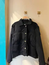 Campera negra con flecos en el pecho y en los hombros, con cuello camisero, cierre frontal con botones a presión y bolsillos de parche.