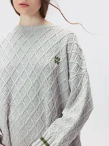 Sweater gris de punto con diseño de rombos, cuello redondo y mangas largas. Cuenta con puños largos acanalados con rayas horizontales en color verde militar y un pequeño logo bordado en el pecho.