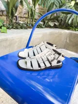 Sandalias planas de cuero color blanco hueso, con múltiples tiras horizontales y hebillas metálicas plateadas. Incluyen pulsera superior desmontable con hebillas laterales para regular el ajuste.