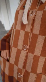 Buzo tipo saco abotonado con diseño de cuadros en tonos marrones y beige, corte recto y oversize. Presenta cuello camisero, cierre frontal con botones personalizados y dos bolsillos delanteros.