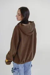 Canguro beige con capucha, cierre frontal parcial y bolsillo tipo canguro. Presenta estampado con texto en el pecho y detalle en la manga.